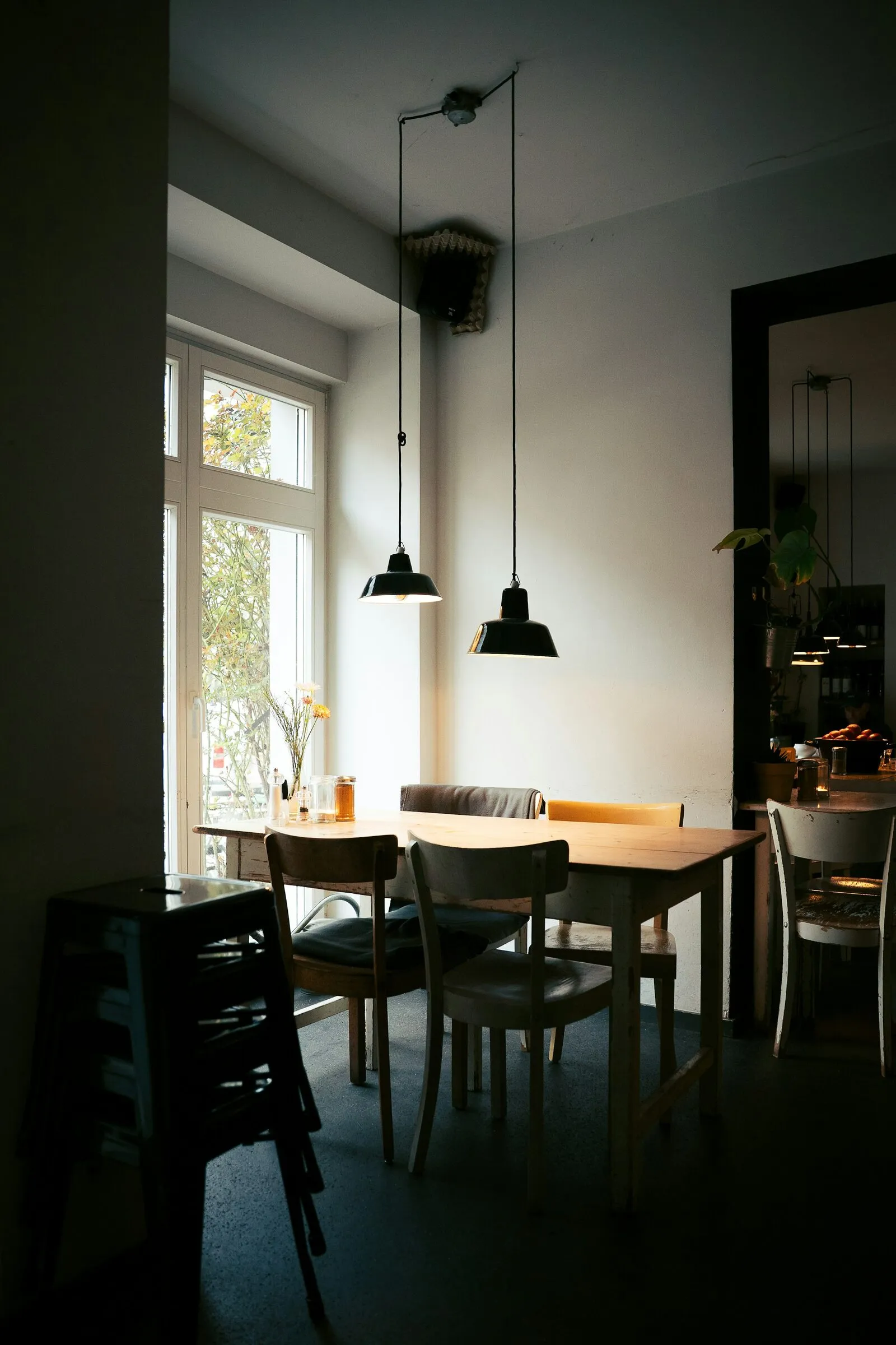 Interier i errët kafeneje me llamba të zeza të varura mbi tavolina druri pranë një dritareje të lartë